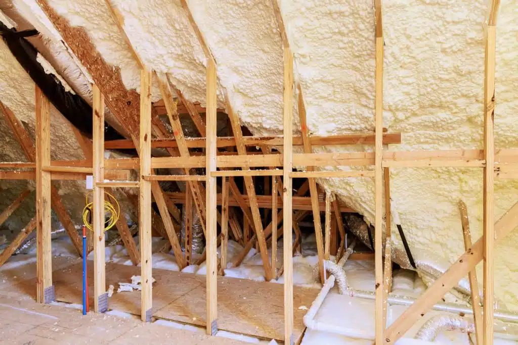 Interior view of a house under construction, showing one side of the upper section just below the roof. The wooden framework is exposed, with spray foam insulation filling the spaces between the beams, preparing the structure for the next phase of construction.